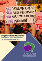 Ao fundo, mulheres em uma manifestação. Em seus cartazes está escrito 